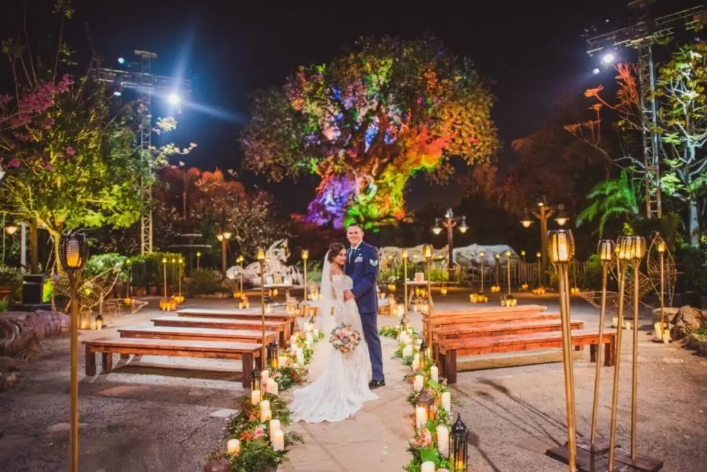 A bride and groom (in military uniform) pose in front of Animal Kingdom's Tree of Life with ceremony seating set up around them.