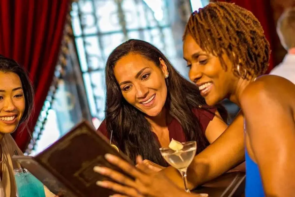 Photo of 3 adult women looking at a menu with cocktails on the table in front of them.