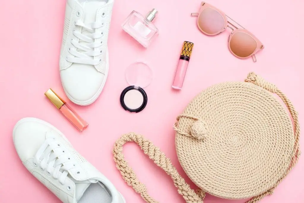 Flat lay photo of a straw purse, white sneakers, pink lip gloss, pink eyeshadow, perfume, and pink sunglasses.