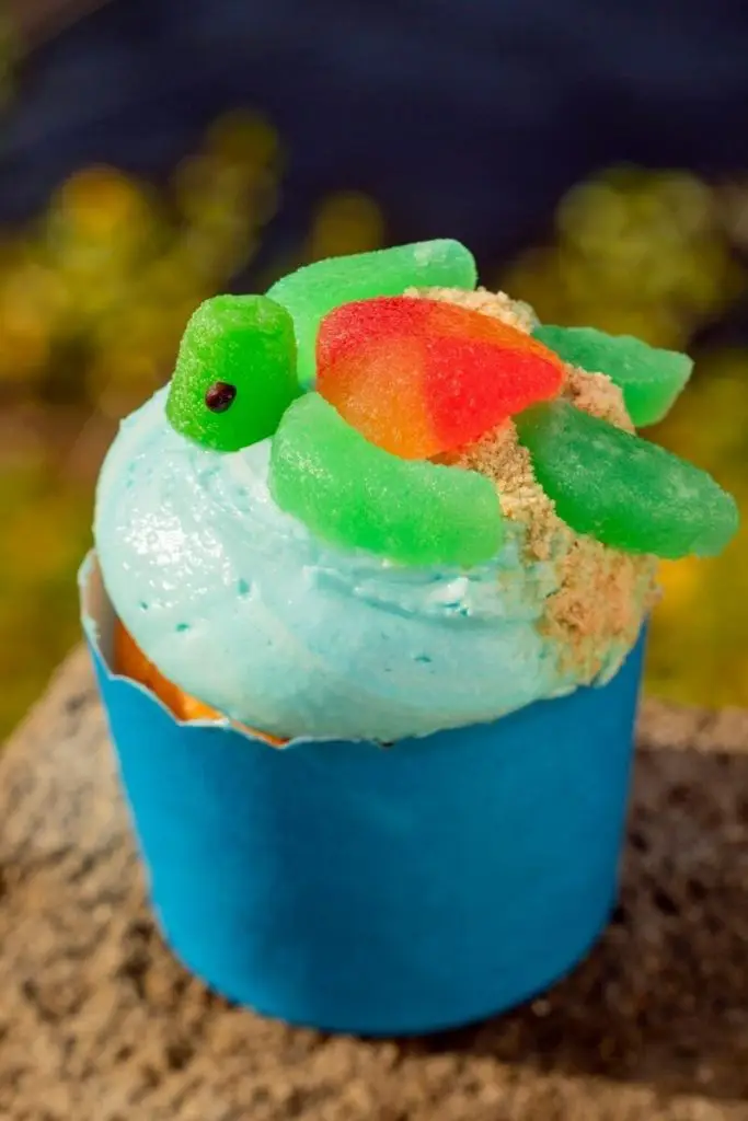 Closeup of a cupcake with blue frosting with a turtle made out of gummy candy and cookie crumb sand on top.