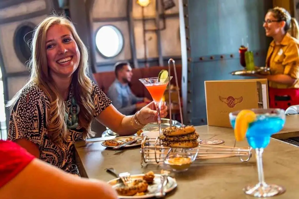 Photo of a woman holding a red cocktail while talking to someone with a server in the background holding a tray of drinks.