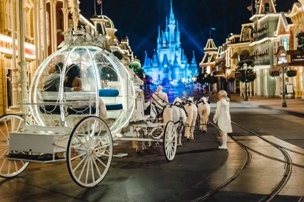 Photo of a bride and groom in a fancy horse-drawn carriage at the start of Main Street in Disney World's Magic Kingdom with Cinderella's castle light up in blue.