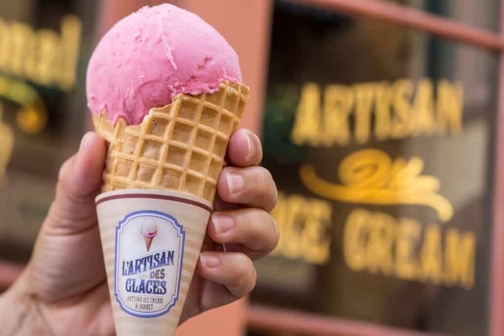 Closeup of a hand holding a waffle cone with raspberry ice cream in front of a store sign.