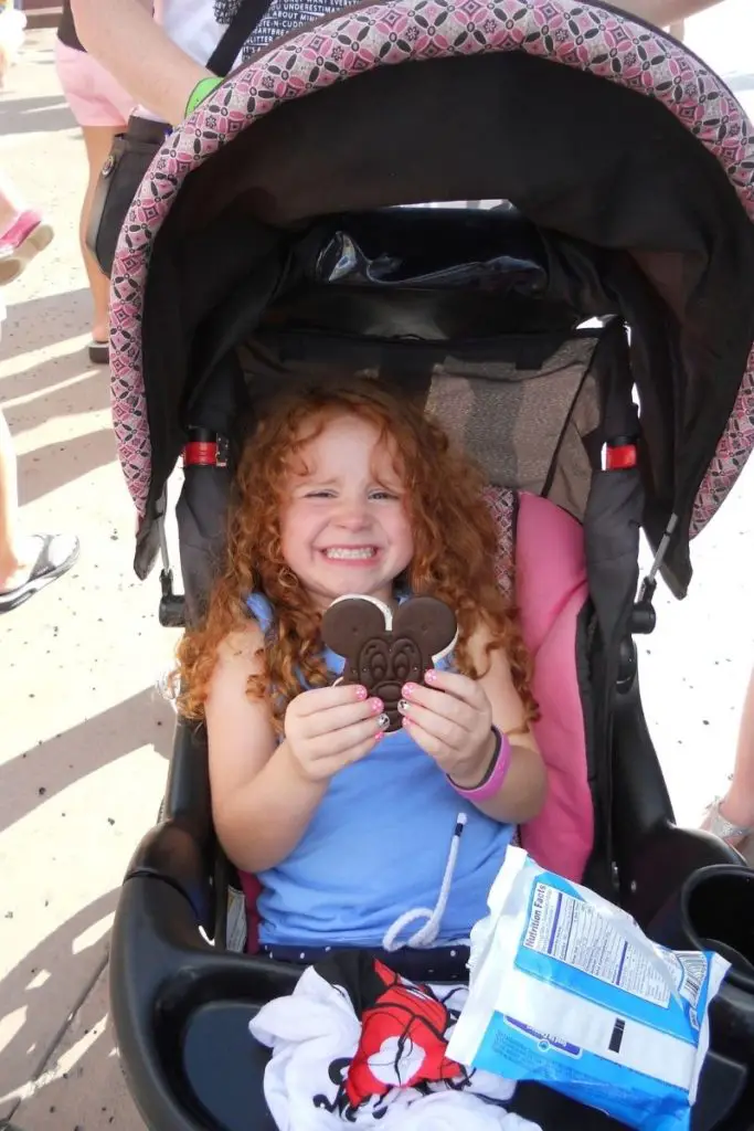 Photo of a young girl smiling in a stroller while holding a Mickey ice cream sandwich, a classic ice cream treat at Disney World.