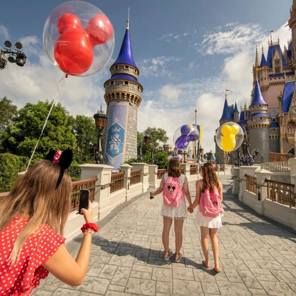 Photo of a young girl taking an Instagram photo of her 2 friends walking toward Cinderella's Castle at Disney World with Mickey balloons in their hands.