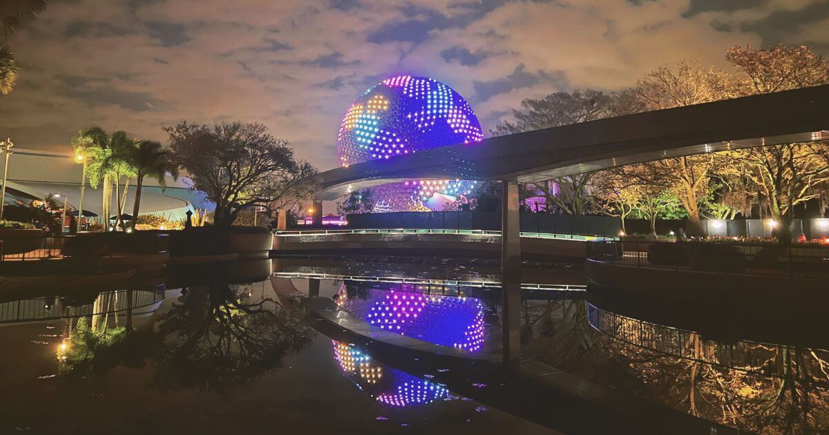 Photo of Spaceship Earth Epcot ride at night lit up with different colors, reflecting in a pond, with the monorail in the foreground.