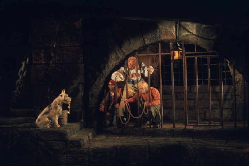 Photo of a scene in Pirates of the Caribbean with 3 pirates in a jail trying to get keys from a dog holding them in his mouth.