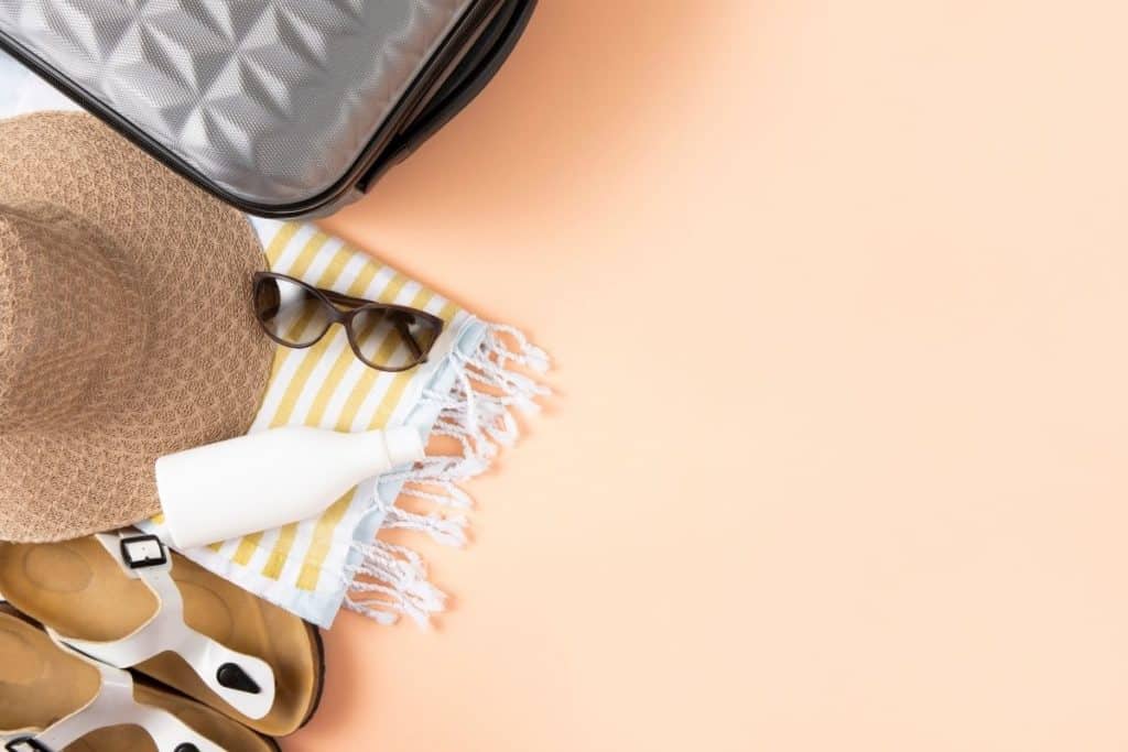 Top-down photo of sandals, beach blanket, sunglasses, and a sun hat next to a suitcase.