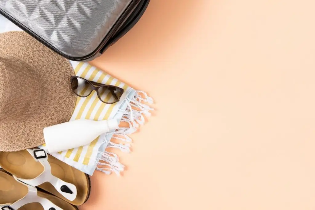 Top-down photo of sandals, beach blanket, sunglasses, and a sun hat next to a suitcase.