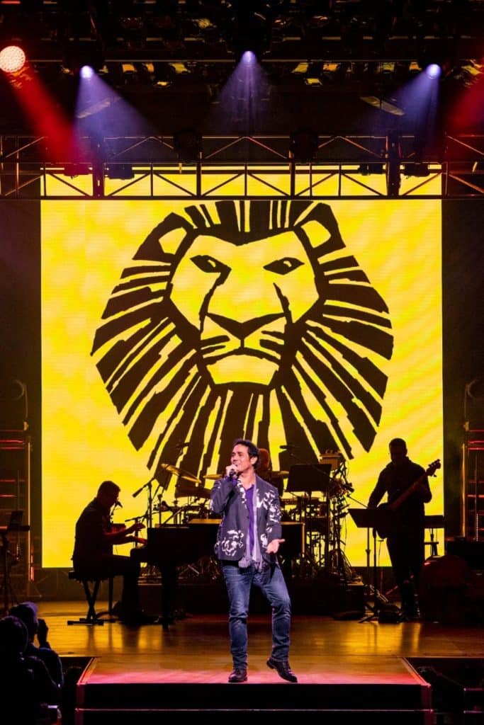 Photo of a man performing on a stage with musicians and a mural from Lion King on Broadway in the background.