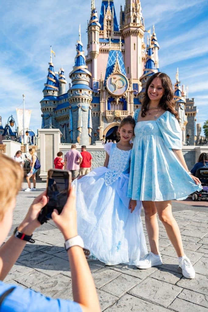 Photo of a mom and daughter posing in front of Cinderella's Castle in fancy dresses while someone takes a photo in the foreground.