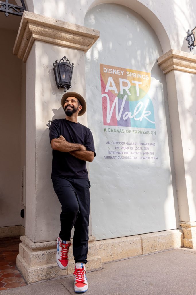 Photo of Brazilian artist Eduardo Kobra posing next to the Disney Springs Art Walk sign.