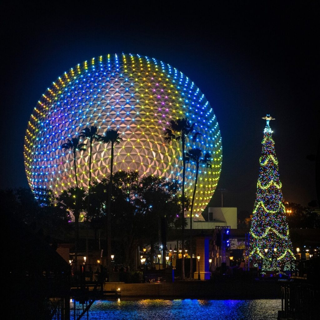Photo of Spaceship Earth at Epcot light up at night with palm trees silhouetted against the ball and a large decorated Christmas tree in the foreground.