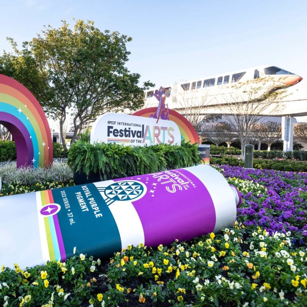 Photo of the Epcot International Festival of the Arts signage in the foreground with the monorail riding by in the background.
