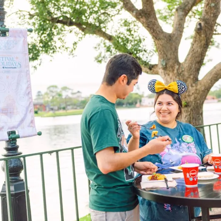 Photo of two young adults eating food at a stand-up table at Epcot.