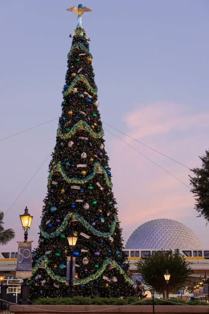 Photo of a Christmas tree with the monorail and Epcot's Spaceship Earth in the background during sunset.
