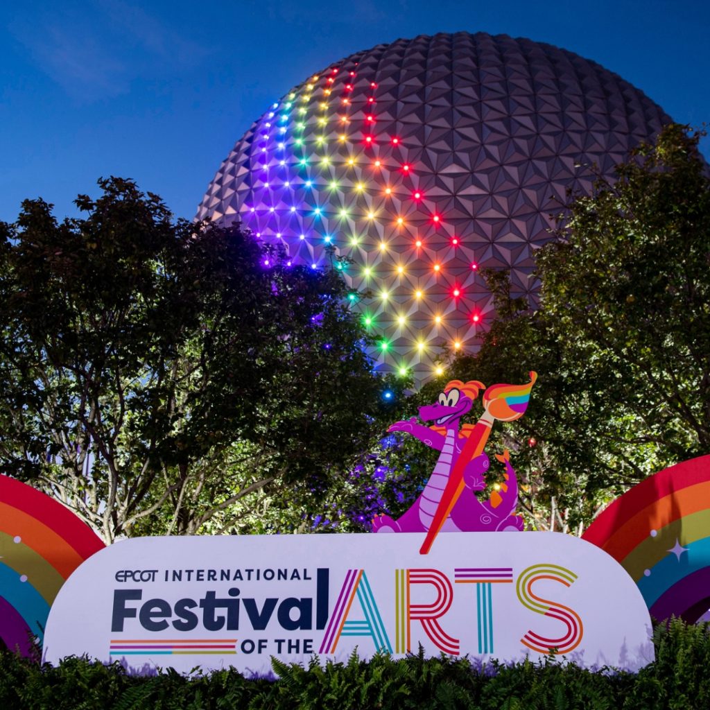 Photo of the Epcot ball with a rainbow light on it with a sign for the Epcot Festival of the Arts in the foreground.
