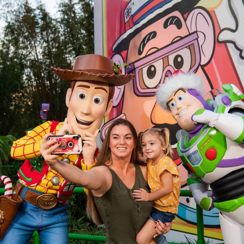 Photo of a woman hold a girl on her hip while taking a selfie with Woody and Buzz Lightyear, both of whom have Christmas outfits on.