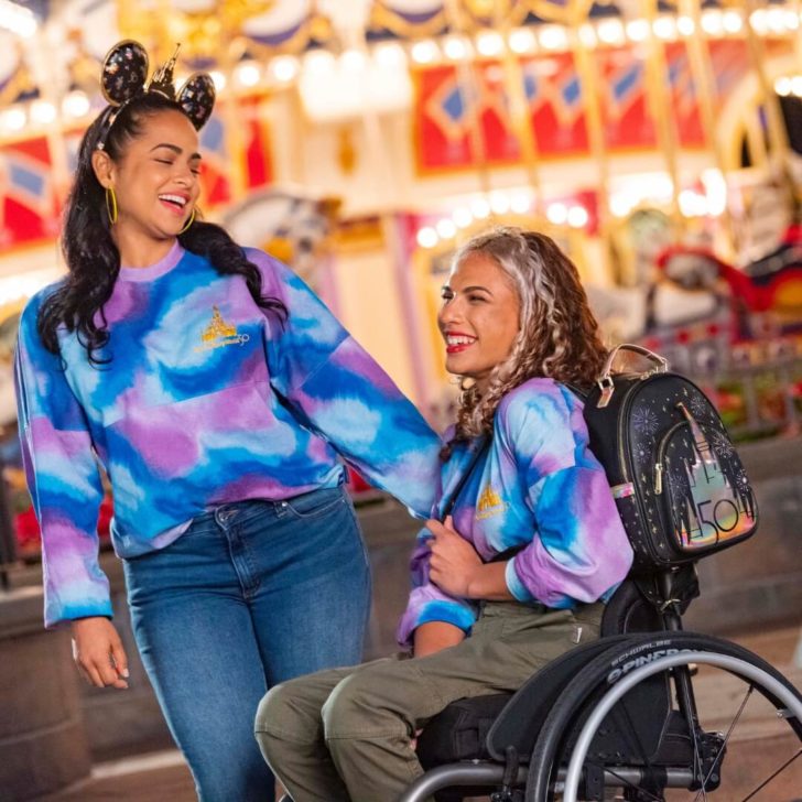 Photo of two young women, one standing and one in a wheelchair, wearing Walt Disney World 50th Anniversary spirit jerseys to keep warm at the park.