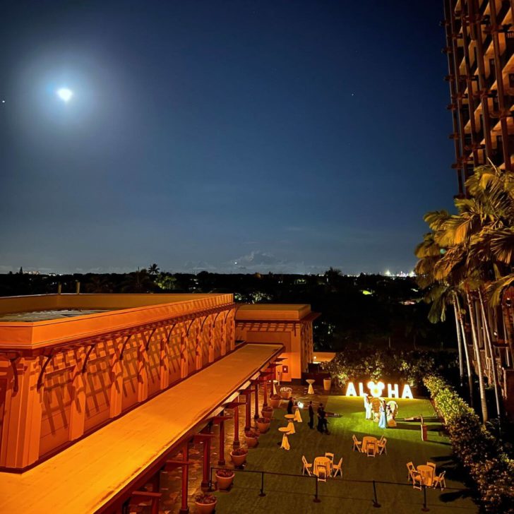 View of the Kipuka Lawn at night from a room balcony at Aulani Resort. In the distance is a lit-up sign that says ALOHA with a Mickey head replacing the O.