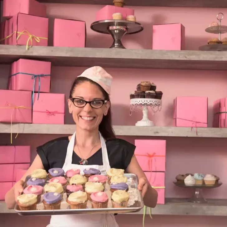 Photo of Erin McKenna posing in her bakery while holding a tray of cupcakes.