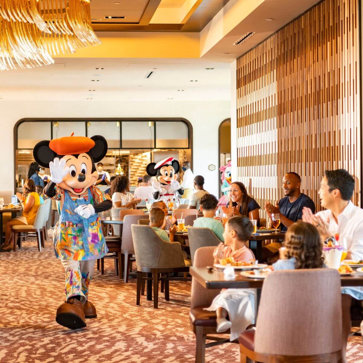 Photo of Mickey Mouse wearing an artist's smock while meeting guests at Topolino's Terrace at Disney's Riviera Resort. Minnie Mouse and Daisy can be seen in the background.