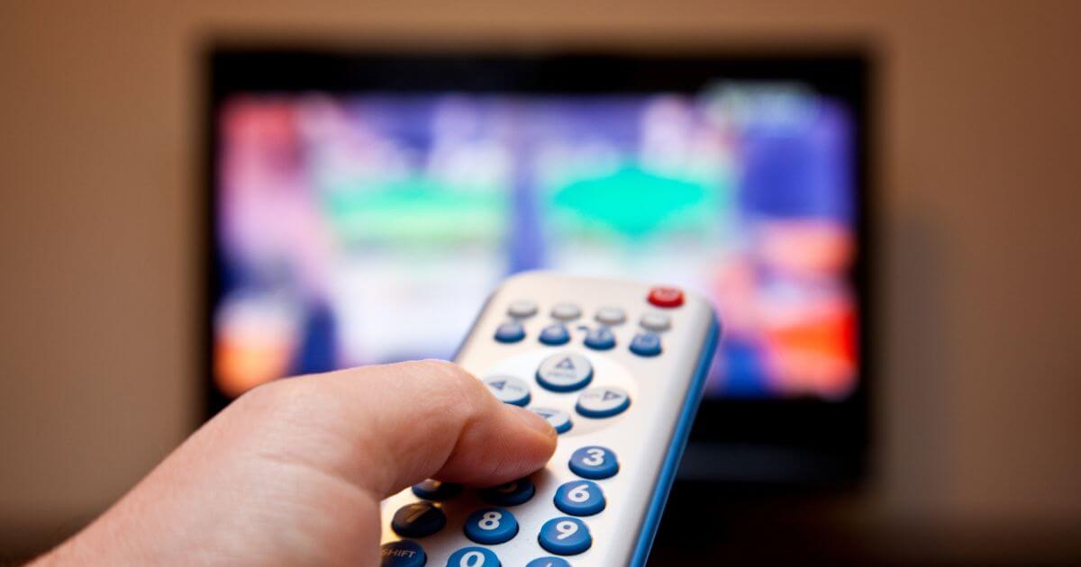 Photo of a hand holding a remote with a blurred TV in the background.