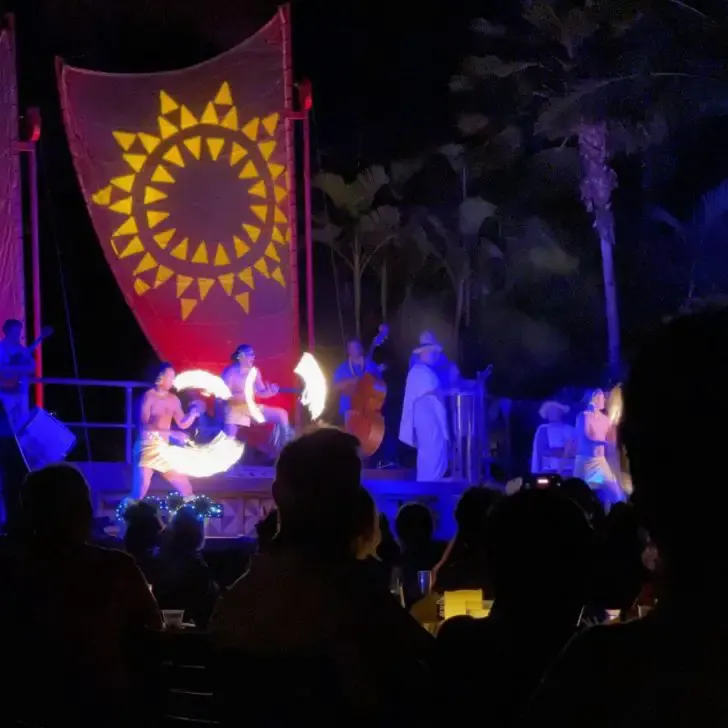 Photo of performers at the Aulani Luau performing Siva Afi, a traditional Samoan fire twirling act with guests in the foreground.