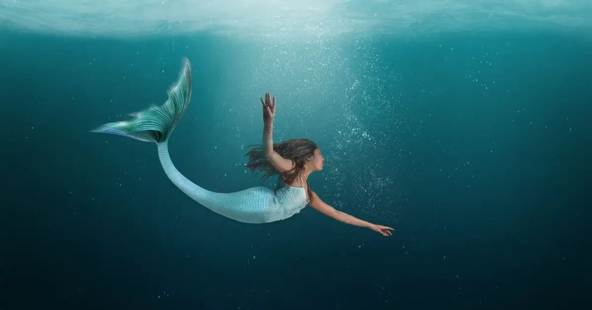 Photo of a mermaid with a light blue and aqua tail and dark hair, swimming under water in the ocean.