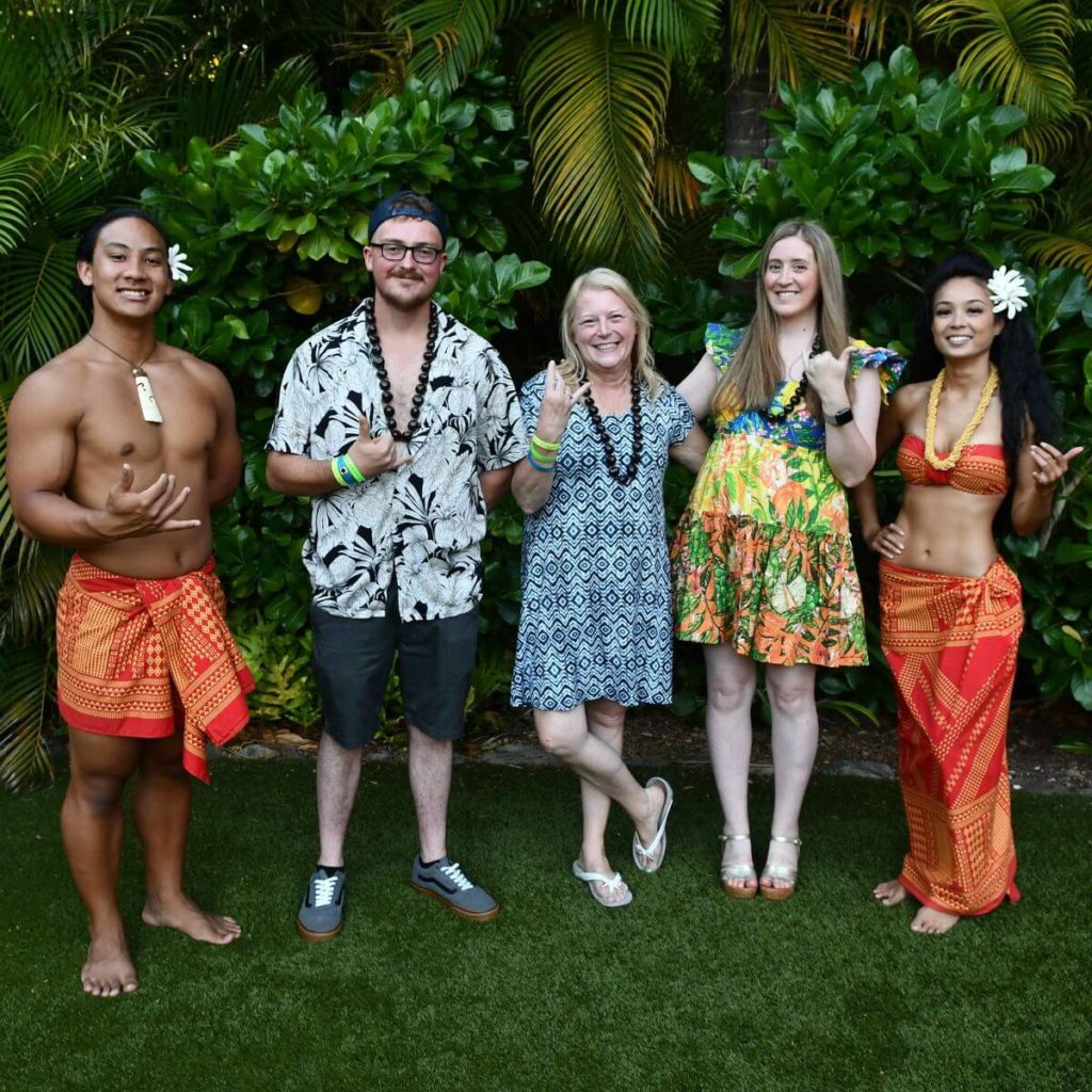 Square photo of a family posing with performers from the Disney Aulani Luau. Everyone is dressed in brightly colored, tropical attire.