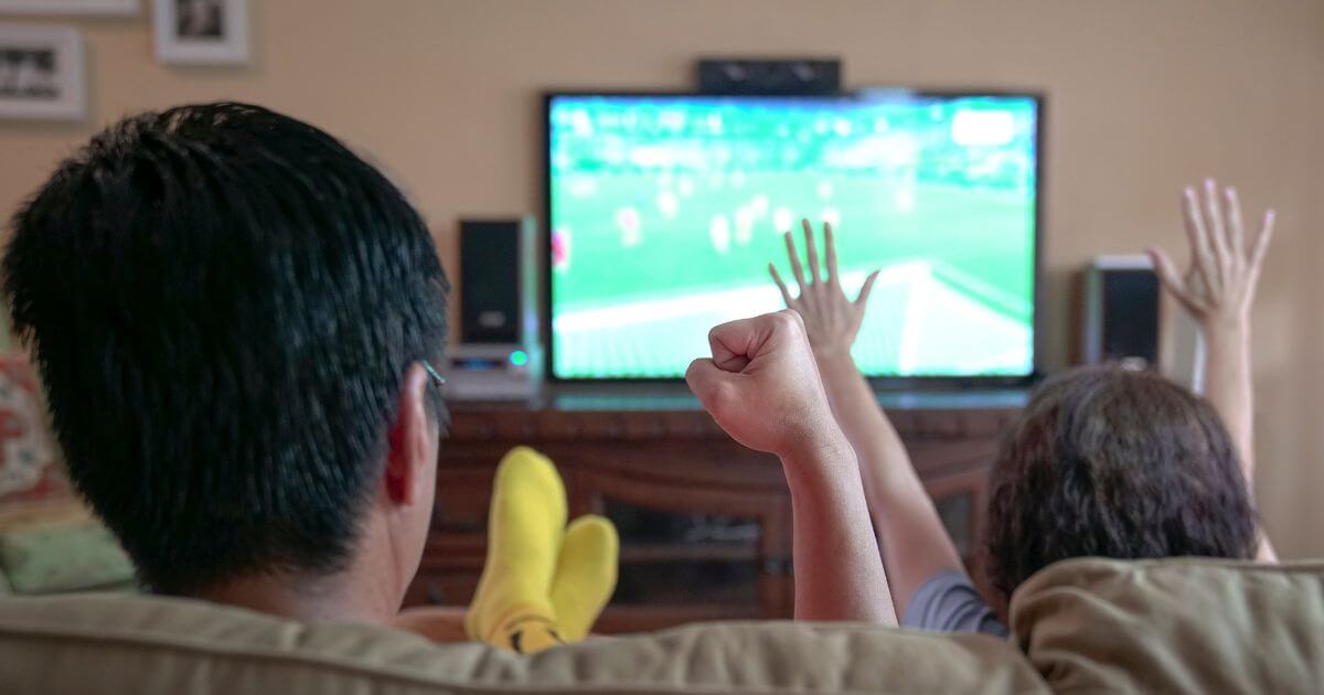 Photo of 2 people sitting on a couch watching a sports game on tv.