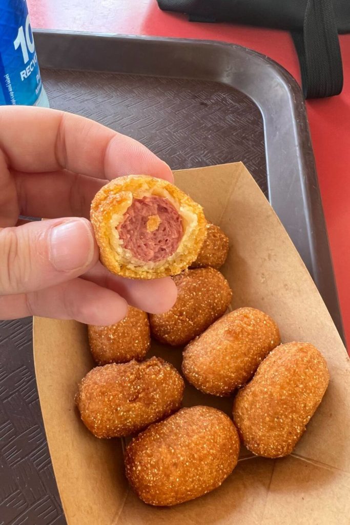 Photo of a basket of corn dog nuggets with a person holding up one that has been half eaten.