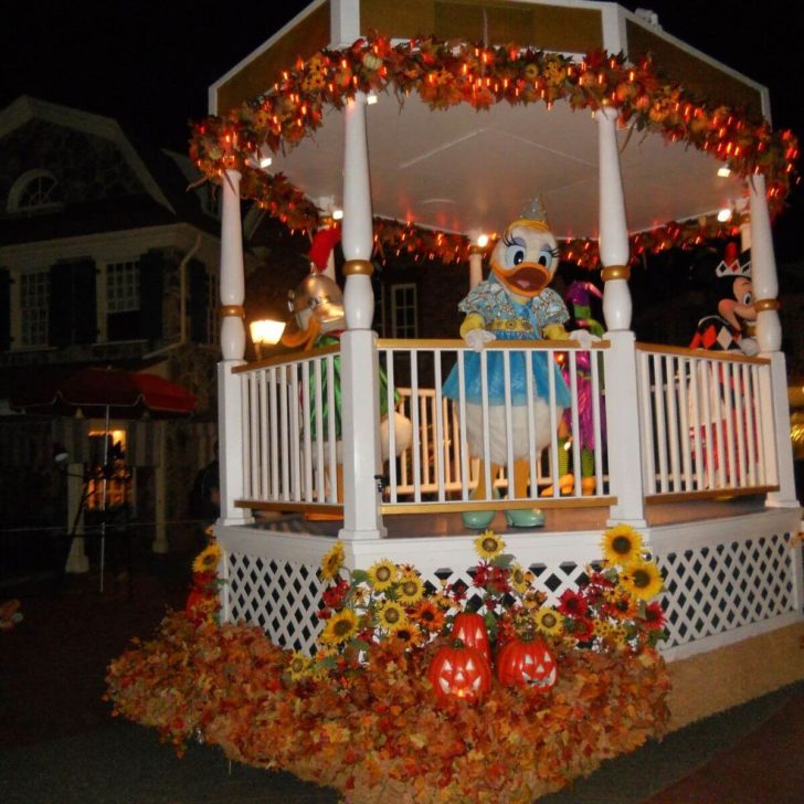 Photo of a floating gazebo decorated with Fall leaves, sunflowers, and pumpkins with Donald, Daisy, Minnie, and Mickey inside, waving to guests.