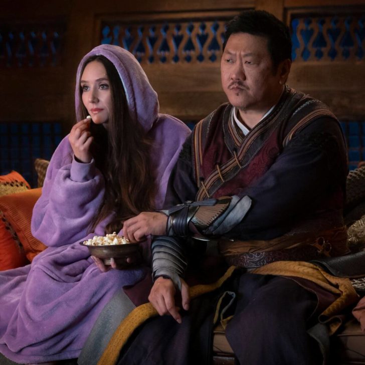 Patty Guggenheim as Madisynn and Benedict Wong as Wong in Marvel Studios' She-Hulk: Attorney At Law. Madisynn is wearing a purple snuggie and Wong is in his usual traditional garb. The two are watching something on TV while eating popcorn.