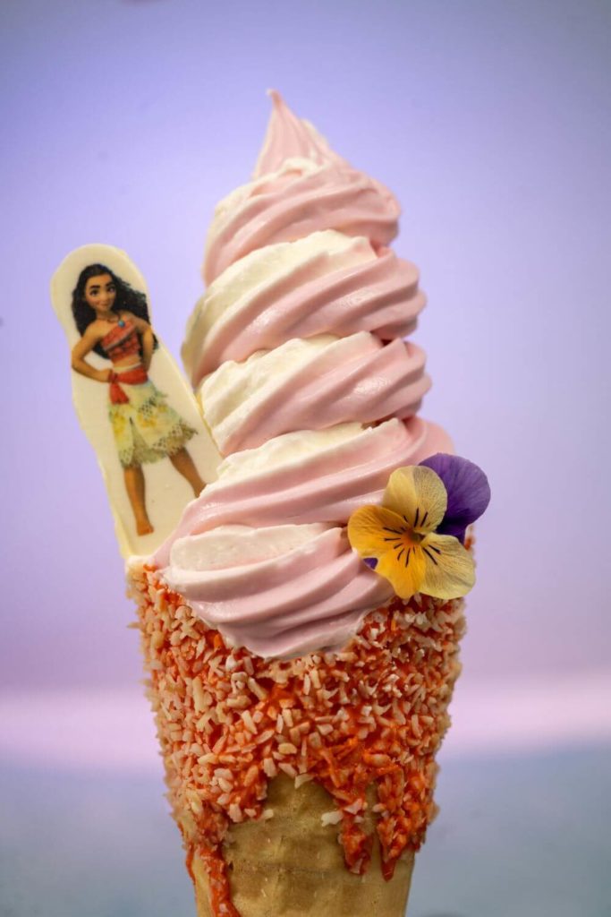 Vertical photo of the Moana Cone from Typhoon Lagoon with strawberry and coconut swirl soft serve in a white chocolate coconut dipped waffle cone and a Moana candy topper.