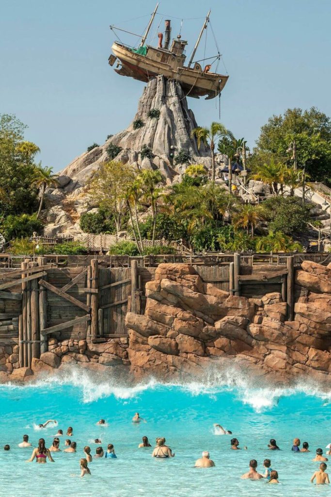 Vertical photo of the wave pool at Disney World's Typhoon Lagoon with lots of people in the pool in the foreground and a shipwrecked boat at the top of a mountain in the back.