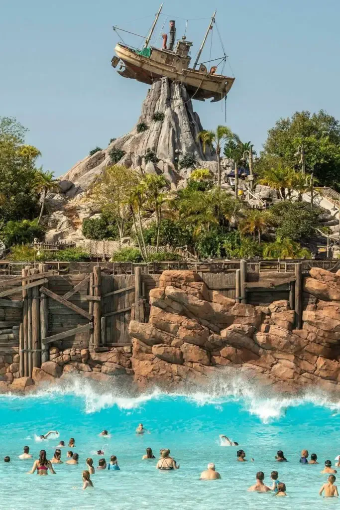 Vertical photo of the wave pool at Disney World's Typhoon Lagoon with lots of people in the pool in the foreground and a shipwrecked boat at the top of a mountain in the back.
