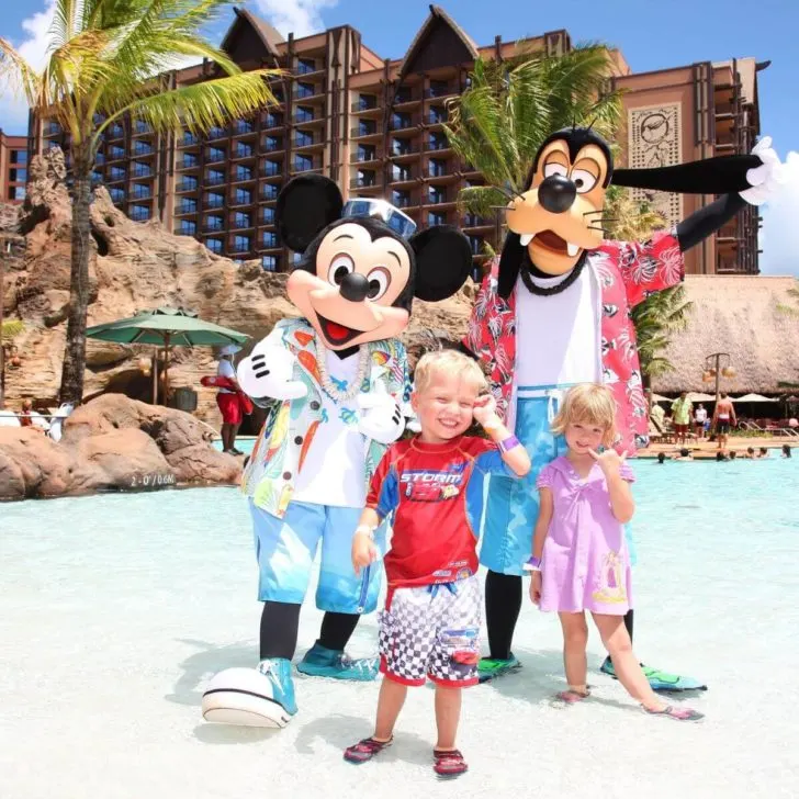 Photo of Mickey Mouse and Goofy in Hawaiian-style pool outfits posing with two young kids at the Waihalohe Pool at Aulani Resort in Hawaii.