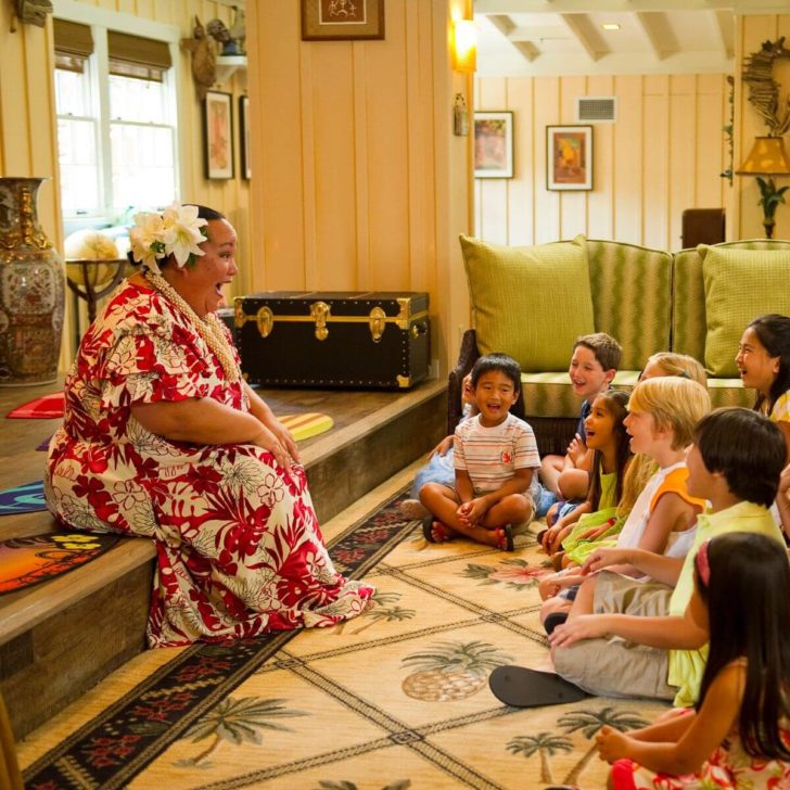 Photo of a woman wearing a red and white mumu, floral lei, and beautiful flowers in her hair, telling a story to young kids seated facing her.