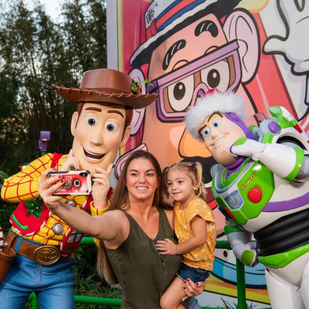 Photo of a woman holding a young girl on her hip, taking a selfie with Buzz and Woody in their Christmas gear.