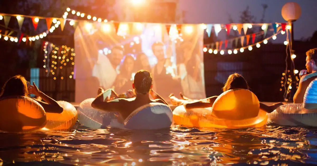 Photo of people sitting in pool floats in a pool with an outdoor movie screen setup in the background.