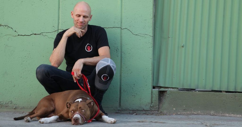 Host of Dog: Impossible, Matt Beisner crouching down on a sidewalk with a dog on a leash laying at his feet.