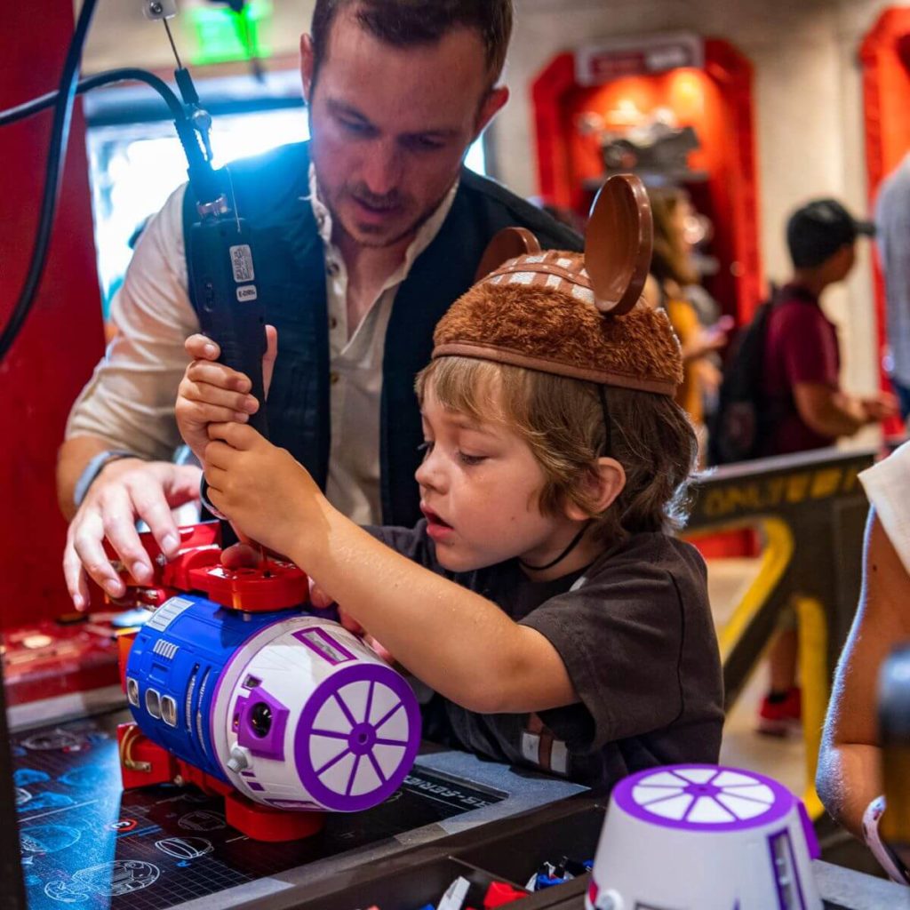 Photo of a young boy building a droid while a male cast member assists.