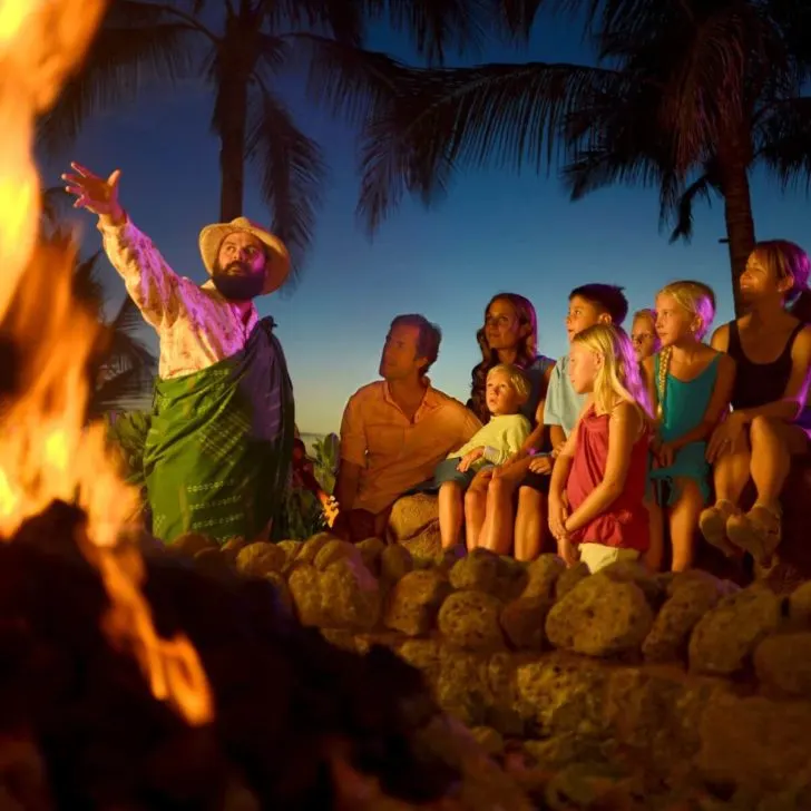 Photo of a man in traditional Hawaiian attire, telling a story to guests nearby a fire pit.