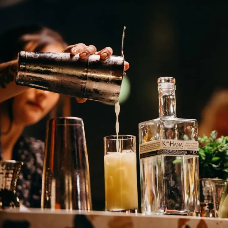 Photo of a woman pouring a cocktail into a glass with a bottle of Kō Hana rum next to it.