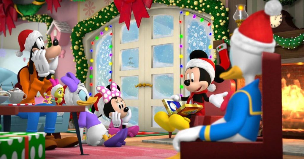 Photo still from the animated Disney Junior movie, Mickey & Minnie Wish Upon a Christmas featuring Goofy, Daisy, Minnie, and Donald listening to Mickey read a story by the fireplace.