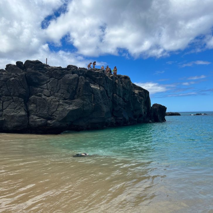 Photo of people standing a large rock in the ocean at Waimea Bay Beach.