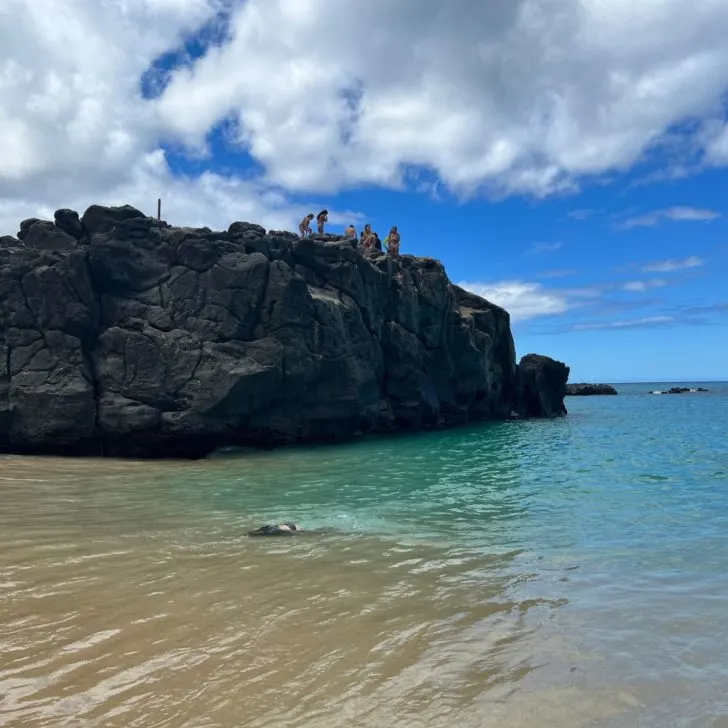 Photo of people standing a large rock in the ocean at Waimea Bay Beach.