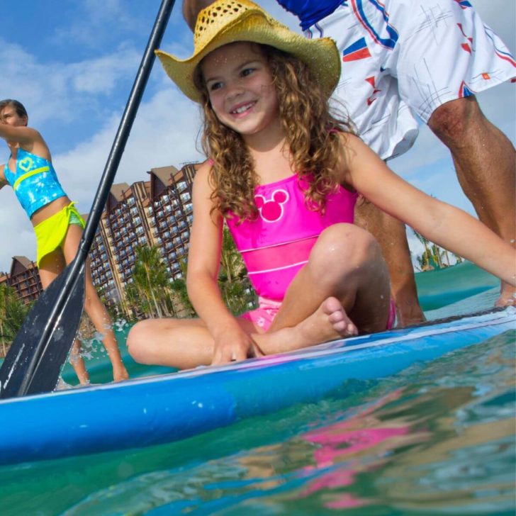 Photo of two people doing stand up paddleboarding in the ocean with a third young girl sitting on one of the paddle boards.