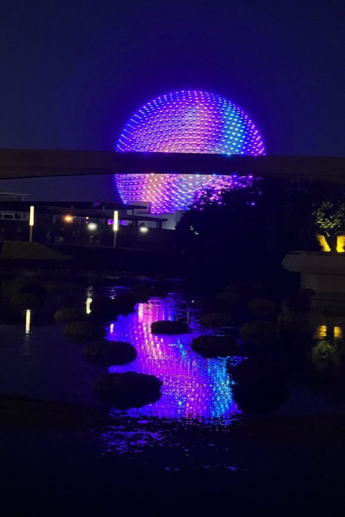 Photo of Spaceship Earth at night, lit up in purple, pink, turquoise, and blue, reflecting in the water in the foreground..